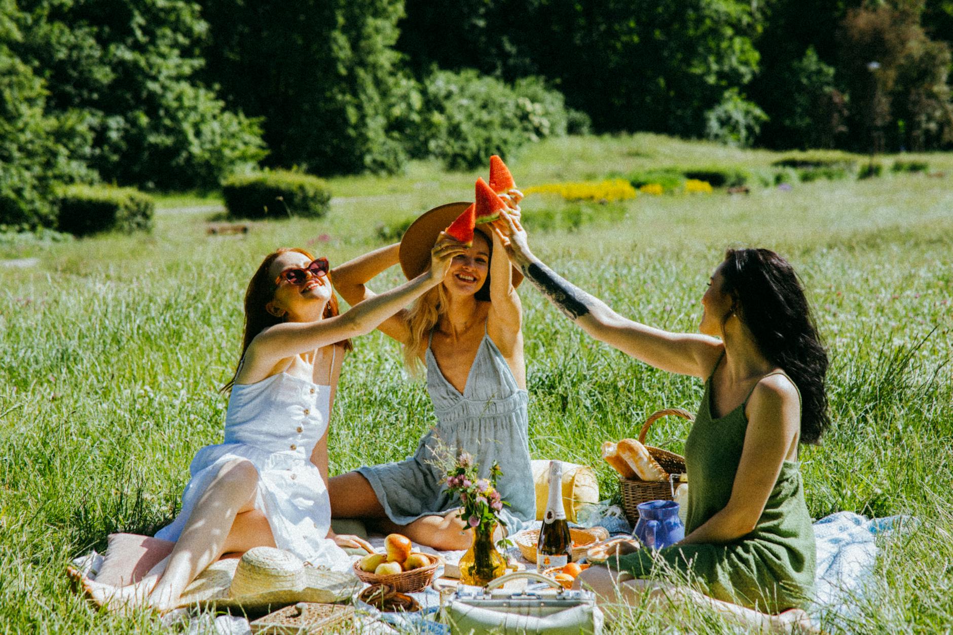 Three women enjoying a picnic in a sunny field, happily holding slices of watermelon and surrounded by food and drinks.